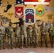 A group of approximately ten U.S. Army soldiers in OCP (Operational Camouflage Pattern) uniforms pose for a formal group photo. Several soldiers are holding trophies, suggesting a recognition or award ceremony. They are standing in front of a large military-themed mural featuring unit insignia, an American flag, military vehicles, and aircraft.