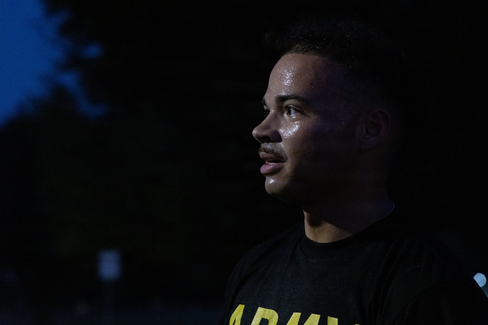 A young man is photographed in profile at night, illuminated by a light source from the right side. He is wearing a black shirt with yellow 'ARMY' lettering visible at the bottom of the frame. His face appears to be perspiring, suggesting recent physical activity.