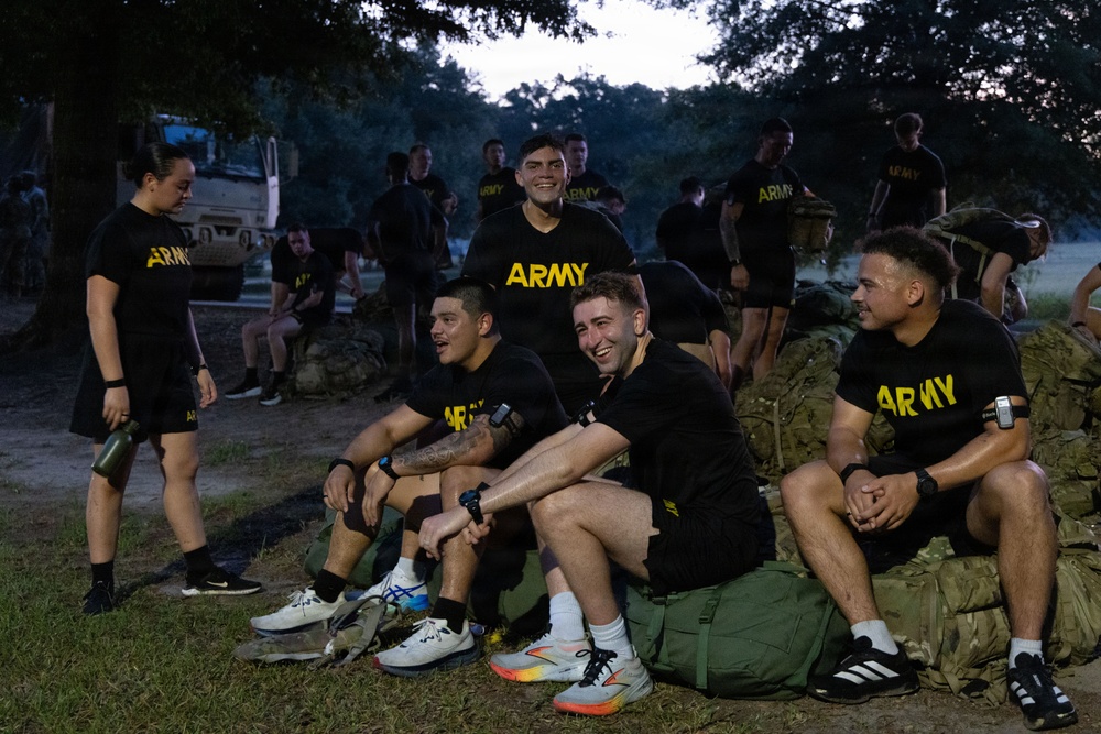 A group of U.S. Army personnel wearing matching black 'ARMY' t-shirts and shorts are gathered outdoors in what appears to be a training or physical fitness setting during early morning or dusk hours. Several soldiers are seated on military duffel bags and rucksacks, laughing and appearing relaxed, suggesting a rest period during or after physical training. A military vehicle is visible in the background among trees.