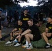 A group of U.S. Army personnel wearing matching black 'ARMY' t-shirts and shorts are gathered outdoors in what appears to be a training or physical fitness setting during early morning or dusk hours. Several soldiers are seated on military duffel bags and rucksacks, laughing and appearing relaxed, suggesting a rest period during or after physical training. A military vehicle is visible in the background among trees.