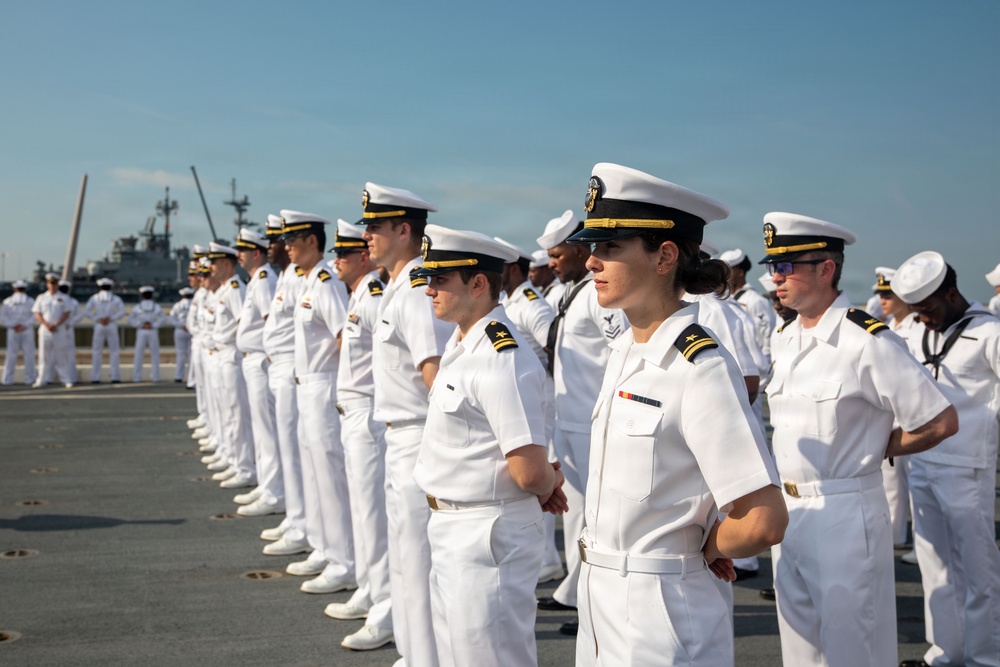 A formation of United States Navy personnel in dress white uniforms are standing at attention on what appears to be a pier or ship deck. The group includes both officers, identifiable by gold insignia and cover braid, and enlisted sailors in standard white uniforms. Naval vessels are visible in the background, suggesting a naval base or port setting.