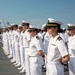 A formation of United States Navy personnel in dress white uniforms are standing at attention on what appears to be a pier or ship deck. The group includes both officers, identifiable by gold insignia and cover braid, and enlisted sailors in standard white uniforms. Naval vessels are visible in the background, suggesting a naval base or port setting.