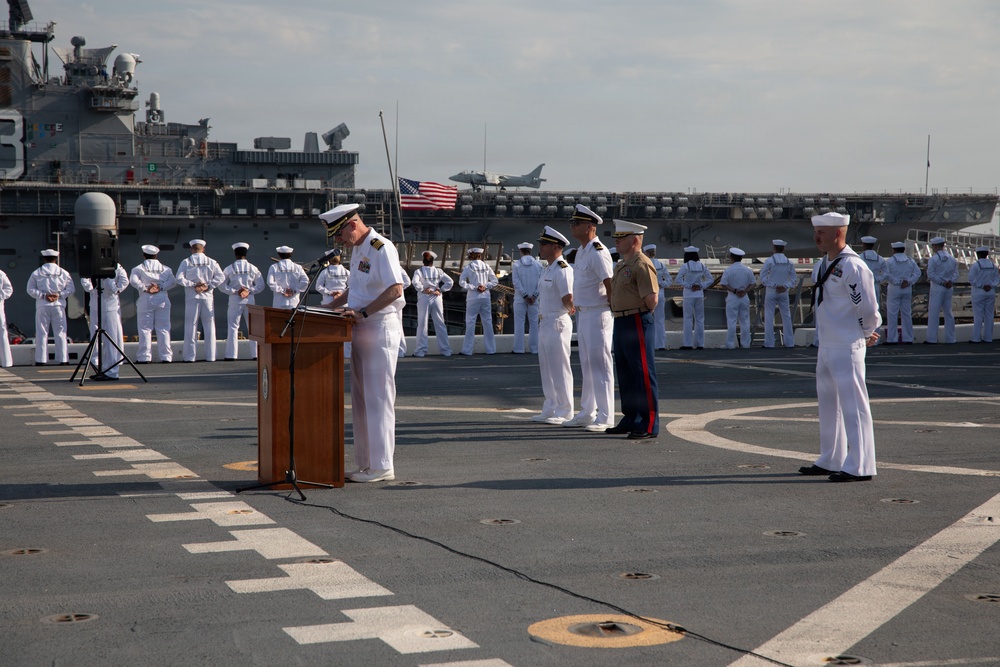 A formal U.S. military ceremony is taking place on the flight deck of a naval vessel, likely a change of command or similar official event. A naval officer in dress whites is speaking at a wooden podium while other officers, including a U.S. Marine in dress blues, stand at attention nearby. Rows of sailors in dress white uniforms are lined up in formation in the background, with a large aircraft carrier and an American flag visible behind them.