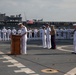 A formal U.S. military ceremony is taking place on the flight deck of a naval vessel, likely a change of command or similar official event. A naval officer in dress whites is speaking at a wooden podium while other officers, including a U.S. Marine in dress blues, stand at attention nearby. Rows of sailors in dress white uniforms are lined up in formation in the background, with a large aircraft carrier and an American flag visible behind them.