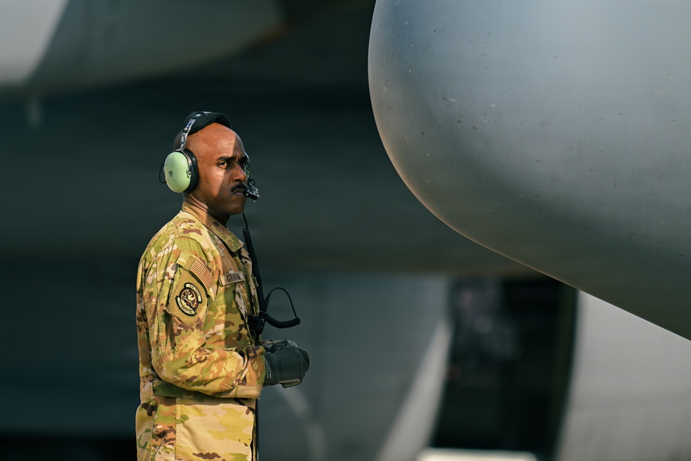 A military service member in OCP (Operational Camouflage Pattern) uniform stands on an aircraft flight line wearing a green aviation headset with boom microphone. The individual appears to be a ground crew member monitoring aircraft operations, positioned near what appears to be a large military aircraft engine nacelle or fuselage. The image is professionally composed with sharp focus on the subject against a blurred aircraft background.
