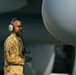 A military service member in OCP (Operational Camouflage Pattern) uniform stands on an aircraft flight line wearing a green aviation headset with boom microphone. The individual appears to be a ground crew member monitoring aircraft operations, positioned near what appears to be a large military aircraft engine nacelle or fuselage. The image is professionally composed with sharp focus on the subject against a blurred aircraft background.