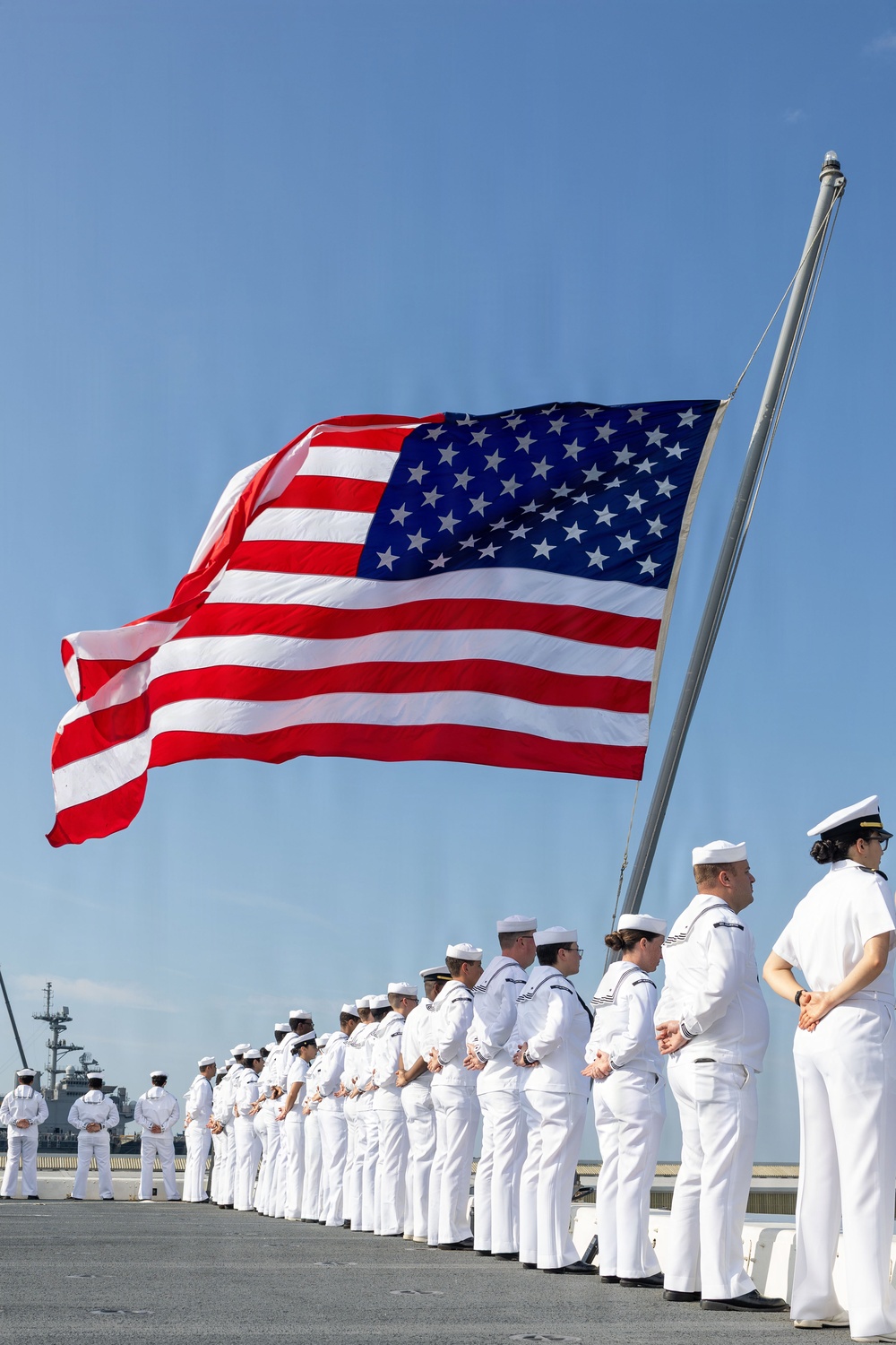 A large formation of U.S. Navy sailors dressed in white service uniforms stand at parade rest on what appears to be a pier or ship deck during a formal ceremony. A large American flag flies prominently from a flagpole in the background against a clear blue sky. Additional naval vessels are visible in the background, suggesting a naval base or port setting.