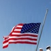 A large formation of U.S. Navy sailors dressed in white service uniforms stand at parade rest on what appears to be a pier or ship deck during a formal ceremony. A large American flag flies prominently from a flagpole in the background against a clear blue sky. Additional naval vessels are visible in the background, suggesting a naval base or port setting.