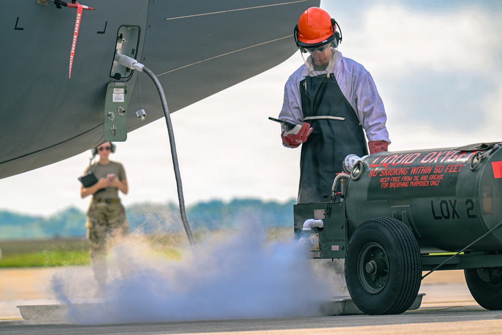 A ground crew technician in full personal protective equipment is servicing a military aircraft with liquid oxygen (LOX) from a mobile LOX cart labeled 'LOX 2'. Visible vapor/fog on the ground indicates active liquid oxygen transfer, which is a cryogenic and hazardous operation. A second personnel member in military uniform stands in the background observing or supervising the procedure.