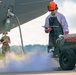 A ground crew technician in full personal protective equipment is servicing a military aircraft with liquid oxygen (LOX) from a mobile LOX cart labeled 'LOX 2'. Visible vapor/fog on the ground indicates active liquid oxygen transfer, which is a cryogenic and hazardous operation. A second personnel member in military uniform stands in the background observing or supervising the procedure.
