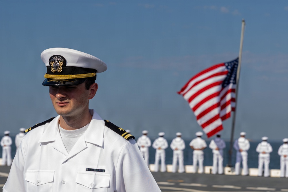 A U.S. Navy officer in white service dress uniform stands in the foreground on what appears to be the deck of a naval vessel. Behind him, a line of sailors in white uniforms stand at attention facing away from the camera, with an American flag flying from a mast in the background. The scene suggests a formal naval ceremony or parade.