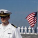 A U.S. Navy officer in white service dress uniform stands in the foreground on what appears to be the deck of a naval vessel. Behind him, a line of sailors in white uniforms stand at attention facing away from the camera, with an American flag flying from a mast in the background. The scene suggests a formal naval ceremony or parade.