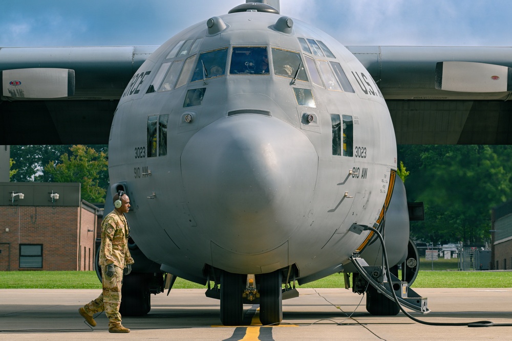 A U.S. Air Force C-130 Hercules transport aircraft, marked with tail number 3023 and assigned to the 910th Airlift Wing (910 AW), is parked on a flight line apron. A ground crew member in camouflage uniform and wearing hearing protection headset is conducting a pre-flight or post-flight walk-around inspection. The aircraft is connected to ground support equipment via cables/hoses on the right side.
