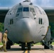 A U.S. Air Force C-130 Hercules transport aircraft, marked with tail number 3023 and assigned to the 910th Airlift Wing (910 AW), is parked on a flight line apron. A ground crew member in camouflage uniform and wearing hearing protection headset is conducting a pre-flight or post-flight walk-around inspection. The aircraft is connected to ground support equipment via cables/hoses on the right side.