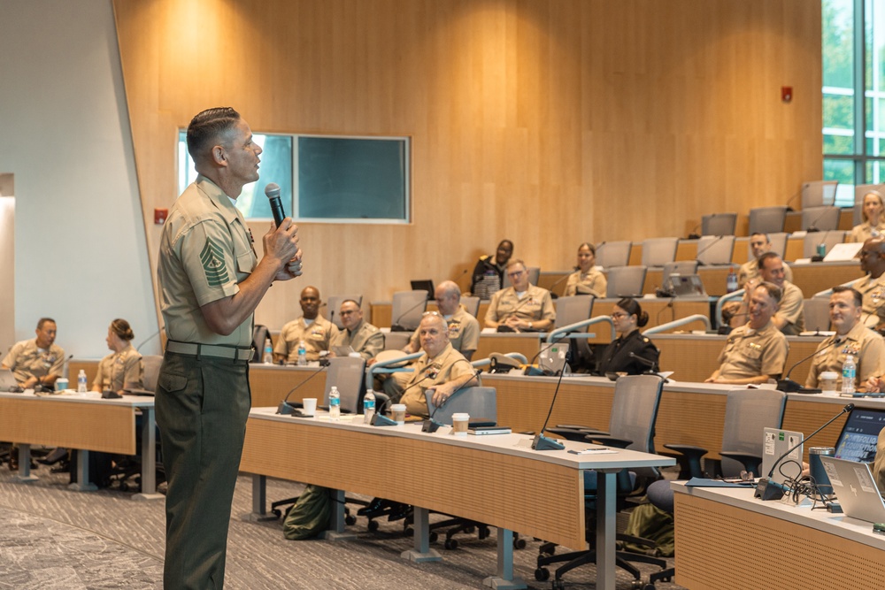 A senior military officer in U.S. Marine Corps service uniform is delivering a presentation or address to an audience of uniformed military personnel in a modern auditorium or lecture hall. The speaker holds a microphone and appears to be engaged in active speech, while the seated audience listens attentively. The setting appears to be a formal military educational or briefing environment.