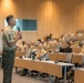 A senior military officer in U.S. Marine Corps service uniform is delivering a presentation or address to an audience of uniformed military personnel in a modern auditorium or lecture hall. The speaker holds a microphone and appears to be engaged in active speech, while the seated audience listens attentively. The setting appears to be a formal military educational or briefing environment.