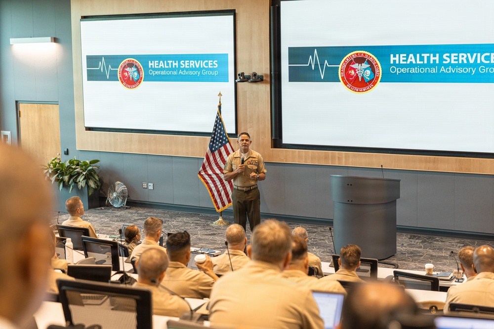A military officer in U.S. Marine Corps uniform is presenting at a formal briefing or advisory group meeting. The presentation screens display 'Health Services Operational Advisory Group' branding, suggesting a medical or health policy discussion. An audience of uniformed military personnel is seated in a tiered conference room setting.