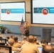A military officer in U.S. Marine Corps uniform is presenting at a formal briefing or advisory group meeting. The presentation screens display 'Health Services Operational Advisory Group' branding, suggesting a medical or health policy discussion. An audience of uniformed military personnel is seated in a tiered conference room setting.