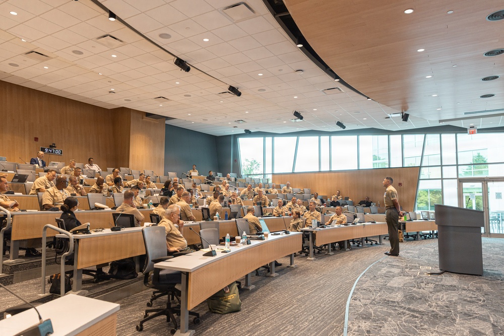 A military briefing or lecture is taking place in a modern auditorium-style conference room. Approximately 50-70 personnel wearing tan/khaki military uniforms are seated at tiered rows of tables, facing a single speaker standing at a podium. The facility appears to be a contemporary military education or command center with wood paneling, recessed lighting, and large windows.