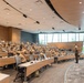A military briefing or lecture is taking place in a modern auditorium-style conference room. Approximately 50-70 personnel wearing tan/khaki military uniforms are seated at tiered rows of tables, facing a single speaker standing at a podium. The facility appears to be a contemporary military education or command center with wood paneling, recessed lighting, and large windows.