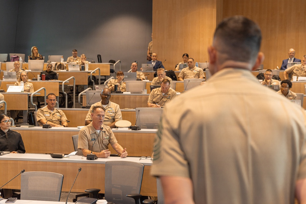 A military briefing or lecture is taking place in a modern tiered auditorium or conference room. The majority of attendees are wearing U.S. Navy service khaki uniforms, with a speaker standing at the front with his back to the camera addressing the seated audience. A uniformed officer in the front row appears to be responding or asking a question at a microphone.