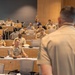 A military briefing or lecture is taking place in a modern tiered auditorium or conference room. The majority of attendees are wearing U.S. Navy service khaki uniforms, with a speaker standing at the front with his back to the camera addressing the seated audience. A uniformed officer in the front row appears to be responding or asking a question at a microphone.