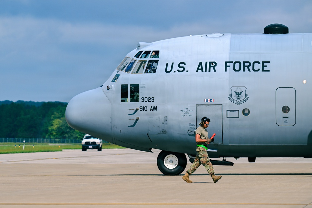 A U.S. Air Force C-130 Hercules transport aircraft, marked with serial number 3023 and assigned to the 910th Airlift Wing (910 AW), is positioned on a tarmac. A ground crew member in military camouflage uniform, wearing hearing protection and carrying signal wands, walks in front of the aircraft's nose gear. The scene appears to be at a military airfield with a white pickup truck visible in the background.