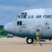 A U.S. Air Force C-130 Hercules transport aircraft, marked with serial number 3023 and assigned to the 910th Airlift Wing (910 AW), is positioned on a tarmac. A ground crew member in military camouflage uniform, wearing hearing protection and carrying signal wands, walks in front of the aircraft's nose gear. The scene appears to be at a military airfield with a white pickup truck visible in the background.