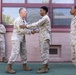 Four U.S. Marines in desert MARPAT camouflage uniforms are gathered outside a building with a pink/mauve exterior. Two Marines in the center are engaged in a handshake while one places a hand on the other's shoulder, suggesting a congratulatory or recognition moment. Two other Marines observe the interaction, one smiling with arms crossed on the left.