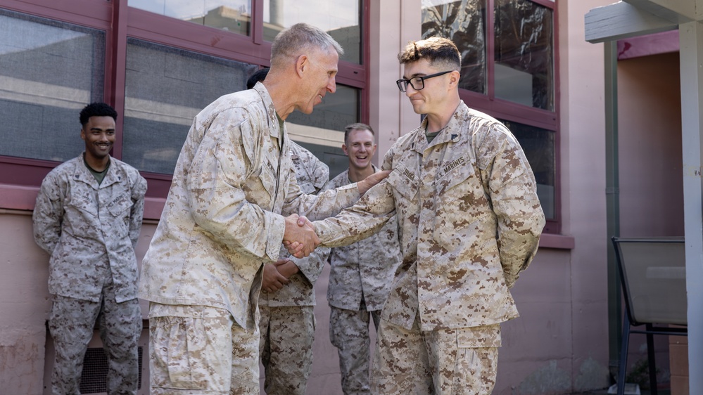 Four U.S. Marines in desert MARPAT camouflage uniforms are gathered outside a building. Two Marines in the foreground are shaking hands, with the senior Marine appearing to congratulate or greet the younger Marine wearing glasses. Two additional Marines stand in the background, smiling and observing the interaction.