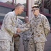 Four U.S. Marines in desert MARPAT camouflage uniforms are gathered outside a building. Two Marines in the foreground are shaking hands, with the senior Marine appearing to congratulate or greet the younger Marine wearing glasses. Two additional Marines stand in the background, smiling and observing the interaction.