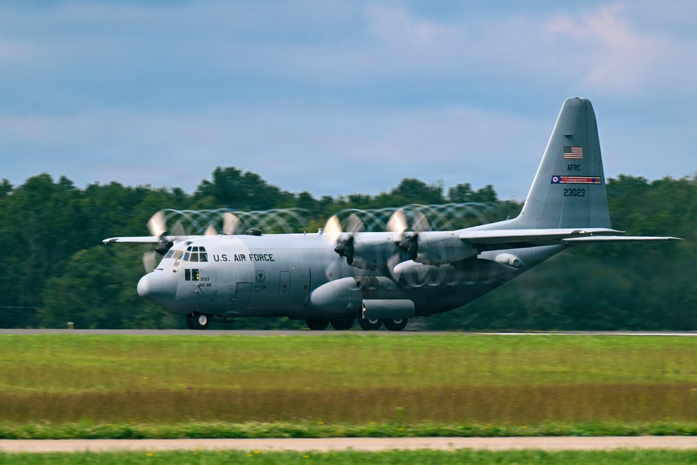 A U.S. Air Force C-130 Hercules military transport aircraft is captured during takeoff or landing roll on a runway. The aircraft bears tail number 23023 and AFRC (Air Force Reserve Command) markings, along with an American flag and what appears to be an Ohio state flag on the tail. The image uses a panning technique, creating motion blur in the background while keeping the aircraft relatively sharp.