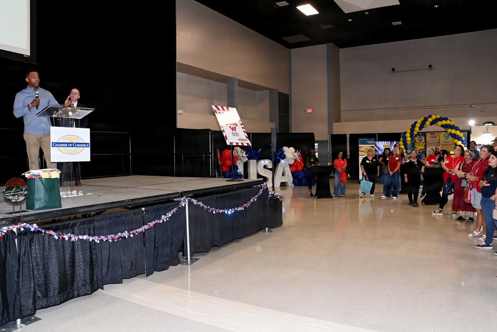 A Chamber of Commerce event is taking place in a large convention hall or event center. A speaker addresses an audience from a raised stage with a podium bearing a Chamber of Commerce logo, while attendees in red and casual attire look on. The venue is decorated with patriotic red, white, and blue balloons, large 'USA' letter props, and a blue and yellow balloon arch.