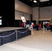 A Chamber of Commerce event is taking place in a large convention hall or event center. A speaker addresses an audience from a raised stage with a podium bearing a Chamber of Commerce logo, while attendees in red and casual attire look on. The venue is decorated with patriotic red, white, and blue balloons, large 'USA' letter props, and a blue and yellow balloon arch.