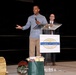A man is speaking at a podium bearing the Yuma County Chamber of Commerce logo at what appears to be a formal event or ceremony. He is holding a microphone and gesturing with his right hand, while two other individuals stand nearby on the stage. Several gift items are visible on the floor in front of the podium.