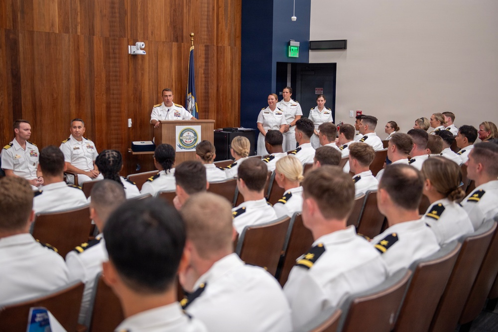 A formal U.S. Navy military ceremony or briefing is taking place in an auditorium-style room. A senior officer in white dress uniform is speaking at a podium bearing a Navy seal/emblem, addressing a large audience of seated naval officers and personnel. Two officers stand at attention behind the speaker near the back of the stage area.