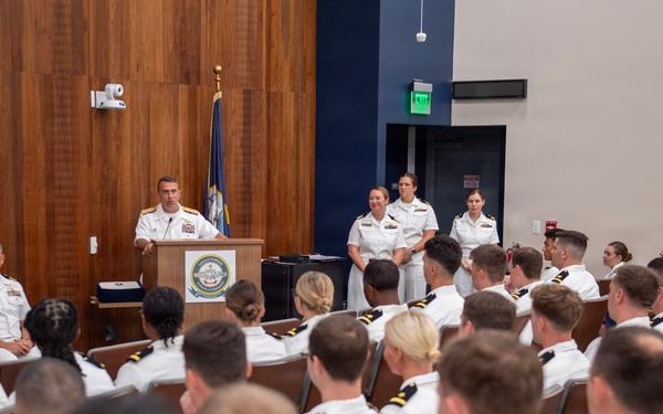 A formal U.S. Navy military ceremony or briefing is taking place in an auditorium-style room. A senior officer in white dress uniform is speaking at a podium bearing a Navy seal/emblem, addressing a large audience of seated naval officers and personnel. Two officers stand at attention behind the speaker near the back of the stage area.