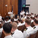 A formal U.S. Navy military ceremony or briefing is taking place in an auditorium-style room. A senior officer in white dress uniform is speaking at a podium bearing a Navy seal/emblem, addressing a large audience of seated naval officers and personnel. Two officers stand at attention behind the speaker near the back of the stage area.