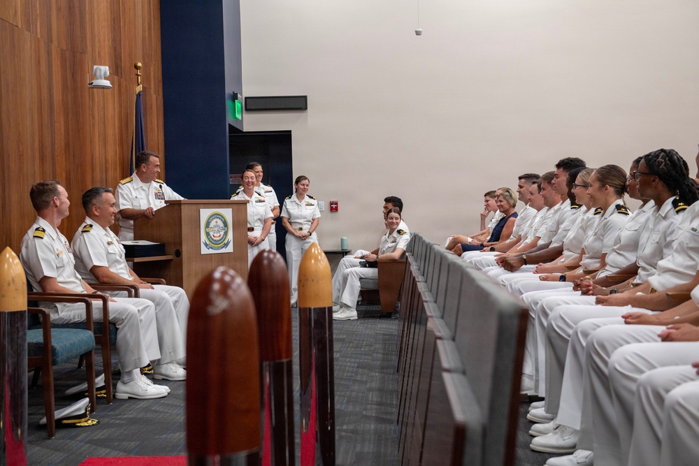 A formal U.S. Navy military ceremony is taking place in an official auditorium or conference room. A senior officer in white dress uniform is speaking at a podium bearing a naval insignia, while rows of uniformed naval personnel are seated and listening attentively. The atmosphere appears positive, with several attendees smiling.