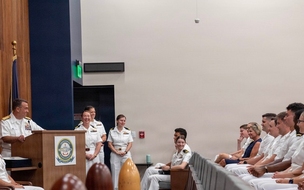 A formal U.S. Navy military ceremony is taking place in an official auditorium or conference room. A senior officer in white dress uniform is speaking at a podium bearing a naval insignia, while rows of uniformed naval personnel are seated and listening attentively. The atmosphere appears positive, with several attendees smiling.