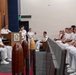 A formal U.S. Navy military ceremony is taking place in an official auditorium or conference room. A senior officer in white dress uniform is speaking at a podium bearing a naval insignia, while rows of uniformed naval personnel are seated and listening attentively. The atmosphere appears positive, with several attendees smiling.