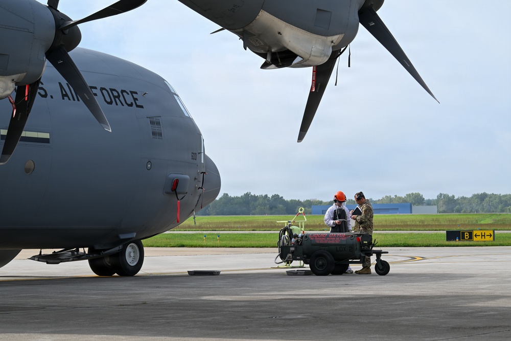 Two personnel are standing beside a U.S. Air Force military transport aircraft on a flight line, reviewing documents or a tablet near ground support equipment. One individual is wearing a red hard hat and white coat, while the other is in military camouflage uniform. A liquid oxygen (LOX) servicing cart is positioned between them and the aircraft.