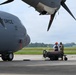 Two personnel are standing beside a U.S. Air Force military transport aircraft on a flight line, reviewing documents or a tablet near ground support equipment. One individual is wearing a red hard hat and white coat, while the other is in military camouflage uniform. A liquid oxygen (LOX) servicing cart is positioned between them and the aircraft.