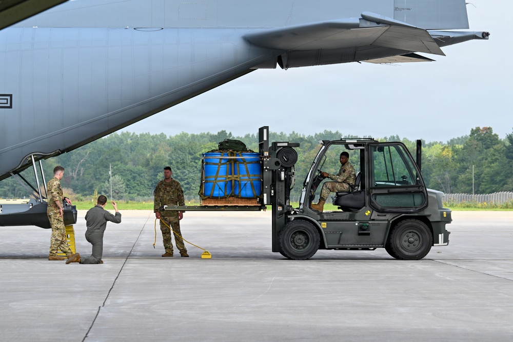 Military personnel are loading cargo onto a large military transport aircraft using a forklift on a concrete tarmac. The cargo consists of blue containers secured with yellow strapping on a pallet. At least four service members in military uniforms are involved in the loading operation.