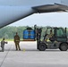 Military personnel are loading cargo onto a large military transport aircraft using a forklift on a concrete tarmac. The cargo consists of blue containers secured with yellow strapping on a pallet. At least four service members in military uniforms are involved in the loading operation.