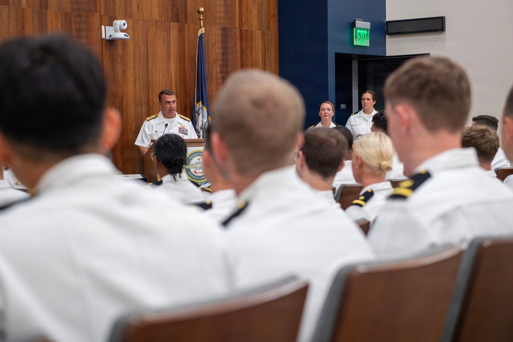 A senior naval officer in white dress uniform is addressing an audience of uniformed naval personnel from a podium in a formal meeting room. Two female officers stand at attention behind and to the side of the speaker. The setting appears to be an official military briefing or ceremony.