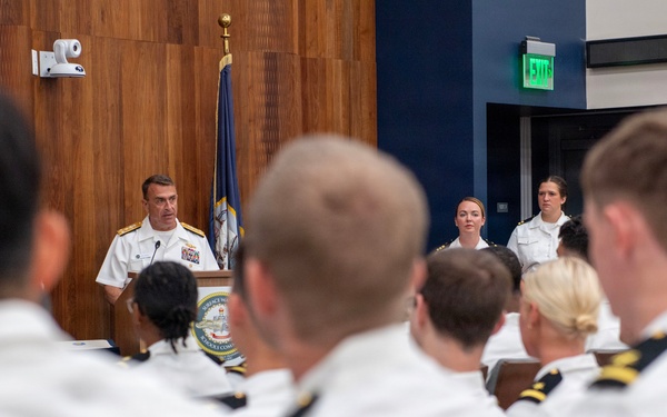 A senior naval officer in white dress uniform is addressing an audience of uniformed naval personnel from a podium in a formal meeting room. Two female officers stand at attention behind and to the side of the speaker. The setting appears to be an official military briefing or ceremony.