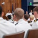A senior naval officer in white dress uniform is addressing an audience of uniformed naval personnel from a podium in a formal meeting room. Two female officers stand at attention behind and to the side of the speaker. The setting appears to be an official military briefing or ceremony.