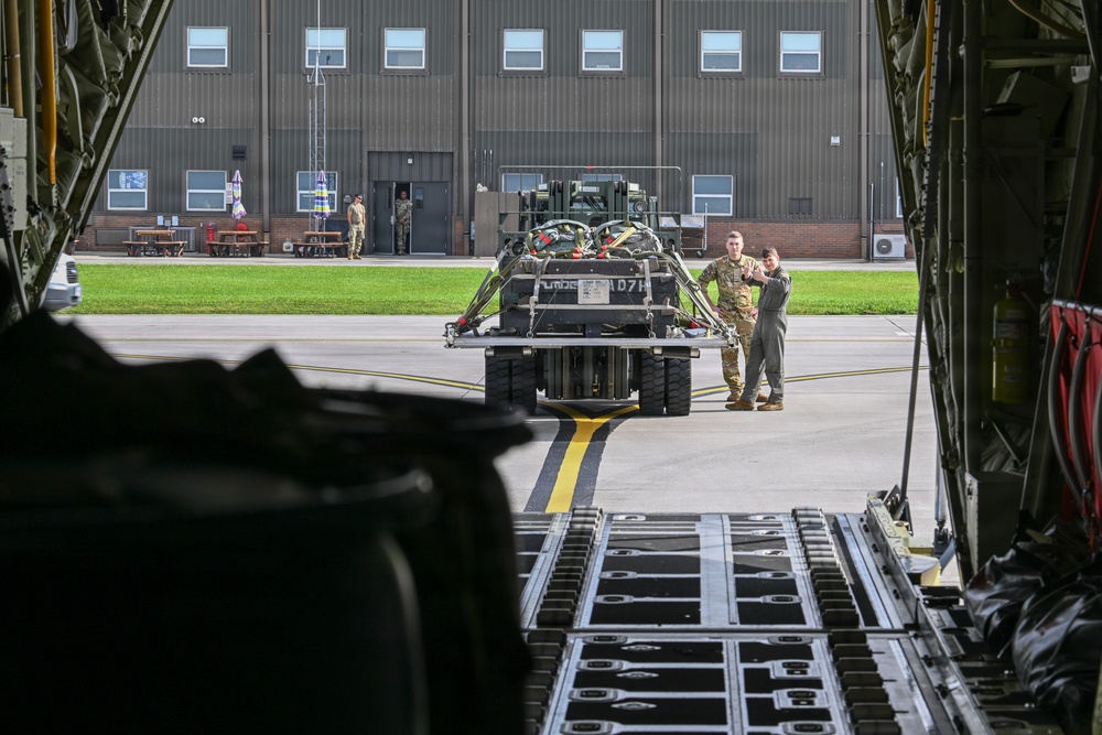 The image is taken from inside a large military cargo aircraft, looking out through the open rear ramp toward a military airfield. Two uniformed personnel are standing near a piece of military equipment or vehicle being prepared for loading onto the aircraft. A large hangar building is visible in the background with additional personnel near its entrance.