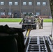 The image is taken from inside a large military cargo aircraft, looking out through the open rear ramp toward a military airfield. Two uniformed personnel are standing near a piece of military equipment or vehicle being prepared for loading onto the aircraft. A large hangar building is visible in the background with additional personnel near its entrance.